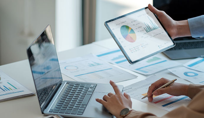 Business professional using a laptop and tablet to review colorful pie charts and financial data reports on a clean office desk